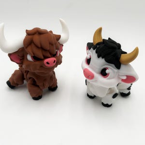 May include: Two stylized bull figurines. One is brown with white horns, pink ears, and red eyes. The other is white with black accents, yellow horns, and pink ears. Both have black hooves and are in a crouching position.