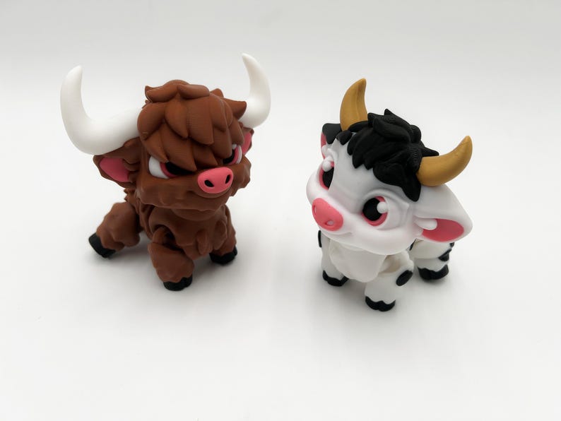 May include: Two stylized bull figurines. One is brown with white horns, pink ears, and red eyes. The other is white with black accents, yellow horns, and pink ears. Both have black hooves and are in a crouching position.