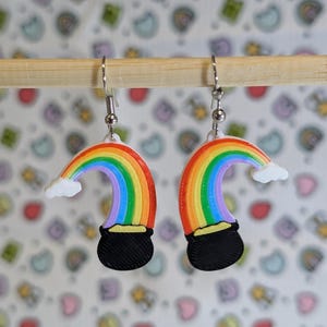 May include: A pair of rainbow and pot of gold earrings. The earrings are made of white and black plastic and feature a rainbow with a cloud and a pot of gold at the end.
