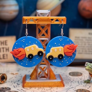 May include: A pair of circular blue earrings featuring a yellow school bus with orange flames trailing behind it. The blue background is decorated with small white dots and thin lines resembling constellations. The earrings hang from silver-toned hooks.