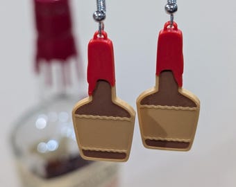 May include: A pair of novelty earrings shaped like miniature whiskey bottles. The earrings feature a red top, brown middle, and tan bottom, with silver-colored ear hooks. The background includes a blurred image of a whiskey bottle.