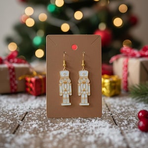 Op de afbeelding: Een paar goudkleurige oorhangers met een wit en goudkleurig notenkrakerontwerp. De oorbellen worden gepresenteerd op een bruine kaart met een rode stip, tegen een feestelijke kerstachtergrond met een boom en ingepakte cadeaus.