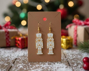 Puede incluir: Un par de pendientes colgantes dorados con un diseño de cascanueces blanco y dorado. Los pendientes se exhiben en una tarjeta marrón con un punto rojo, sobre un fondo navideño festivo con un árbol y regalos envueltos.