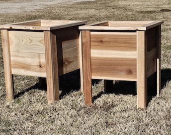 Könnte beinhalten: Zwei Holzkasten mit einer natürlichen Holzoptik. Die Kästen sind quadratisch und haben einen erhöhten Rand oben. Sie stehen auf einer grasbewachsenen Fläche.
