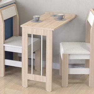 Peut inclure: Une table pliante en bois clair avec deux chaises. La table a un motif à lattes blanches sur le devant et les chaises ont des sièges blancs. La table est dressée avec deux tasses et soucoupes.