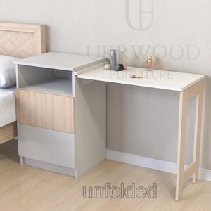 Pode incluir: Uma mesa de cabeceira branca e de madeira com uma mesa de escritório dobrável. A mesa tem uma prateleira e duas gavetas. A mesa de escritório está desdobrada e tem uma pequena garrafa de vidro e artigos de maquiagem. O texto "UKRWOOD FURNITURE" é visível no fundo. O texto "unfolded" é visível em primeiro plano.
