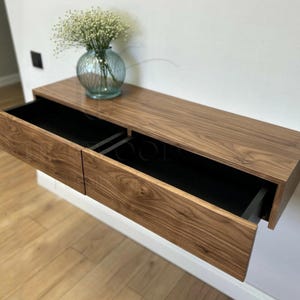 Op de afbeelding: Een wandbevestigde zwevende plank met twee laden. De plank is gemaakt van hout met een natuurlijke afwerking. De laden zijn open en hebben zwarte interieurs. Een vaas met witte bloemen staat op de plank.