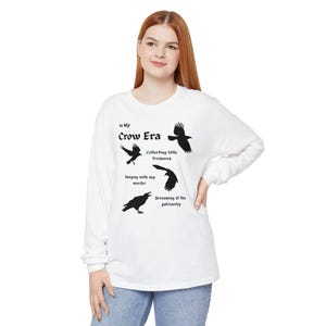 May include: White long-sleeve shirt with a graphic of four black crows and the text "In My Crow Era." The shirt also includes the phrases "Collecting little treasures," "Hanging with my murder," and "Screaming at the patriarchy."