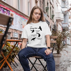 Può includere: T-shirt bianca con la scritta "In My Crow Era" e illustrazioni di corvi. La maglietta presenta frasi come "Collecting little treasures" e "Screaming at the patriarchy."