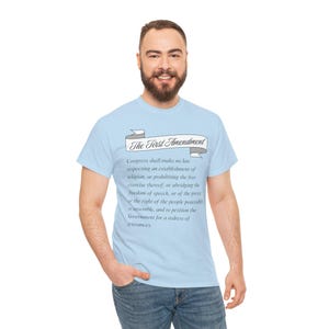 May include: Pale blue t-shirt with the text "The First Amendment" in a banner, and the amendment's text. The shirt is worn by a person in blue jeans.