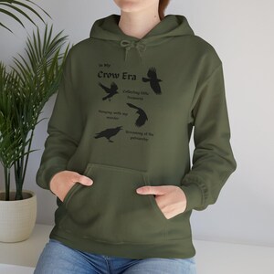 Può includere: Felpa con cappuccio verde oliva con tasca frontale e coulisse. La grafica sul davanti presenta illustrazioni nere di corvi e il testo "In My Crow Era", insieme a frasi come "Collecting little treasures" e "Screaming at the patriarchy."