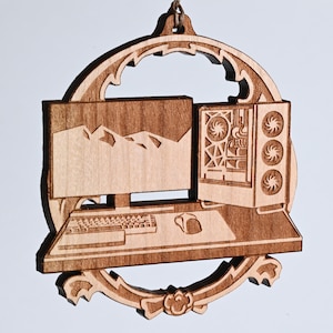 May include: A wooden ornament featuring a computer setup, including a monitor displaying mountains, a keyboard, mouse, and a computer tower with fans. The ornament is framed by an ornate, decorative border and is suspended by a string.