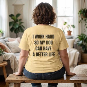Könnte beinhalten: Ein hellgelbes T-Shirt mit dem schwarzen Text "I WORK HARD SO MY DOG CAN HAVE A BETTER LIFE". Die Person trägt eine blaue Jeans und sitzt auf einer Holzbank. Der Hintergrund umfasst eine Couch, Pflanzen und ein Fenster.