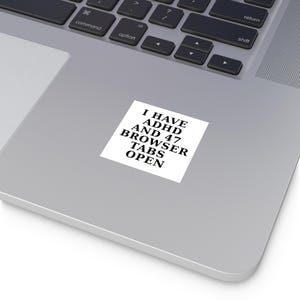 Könnte beinhalten: Ein weißer Aufkleber mit schwarzem Text, der lautet: "I HAVE ADHD AND 47 BROWSER TABS OPEN". Der Aufkleber ist rechteckig und befindet sich auf einer grauen Laptop-Oberfläche. Die Laptop-Tastatur ist im Hintergrund sichtbar.