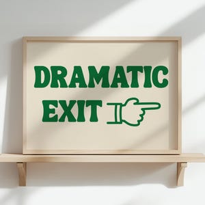 May include: A framed print with the words "DRAMATIC EXIT" in green, with a pointing hand graphic. The artwork is displayed on a light-colored shelf against a white wall.