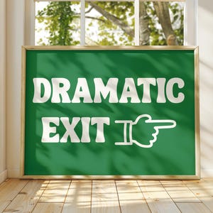 May include: A framed green sign with the words "DRAMATIC EXIT" in white, with a pointing hand graphic. The sign has a gold frame and is displayed indoors near a window.