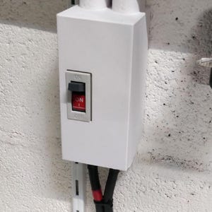 Peut inclure: Un boîtier de disjoncteur électrique blanc avec un interrupteur rouge et noir indiquant "ON" et "OFF". Des câbles noirs avec des rayures rouges sortent du bas. Le boîtier est monté sur un mur blanc texturé.