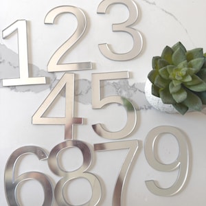 以下が含まれることがあります： 1、2、3、4、5、6、7、8、9の数字がグリッド状に並んだ、シルバーの鏡面仕上げのハウスナンバーセット。各数字は、反射面を持つクリーンなサンセリフ体でデザインされています。右側には小さな緑色の多肉植物が配置されています。