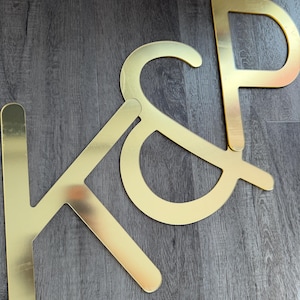 Puede incluir: Un letrero de acrílico de color dorado que presenta las letras K y P conectadas por un símbolo de ampersand. Las letras tienen una superficie lisa y reflectante con un estilo de fuente sans-serif. El letrero consta de tres piezas planas.