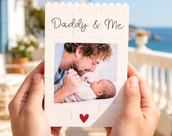 May include: A white brick-style photo frame with the text "Daddy & Me" and a photo of a father and baby. A red heart is at the bottom. The frame is held in front of a scenic background of a house and the ocean.