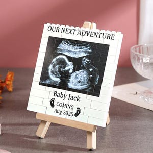 Könnte beinhalten: Ein kleiner Holzstaffelei zeigt ein weißes Schild mit einem Ultraschallbild. Auf dem Schild steht "OUR NEXT ADVENTURE", "Baby Jack COMING Aug 2025" und es sind Babyfüße abgebildet. Der Hintergrund umfasst eine rosa Wand, Blumen und eine Glasschale.