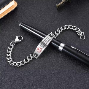 May include: A silver-colored medical ID bracelet with a chain link design. The rectangular tag displays the medical symbol and text: COUMADIN/WARFARIN, ALLERGIES: PENICILLIN, ICETOM G 666.555.5678. The bracelet is resting on a black pen.