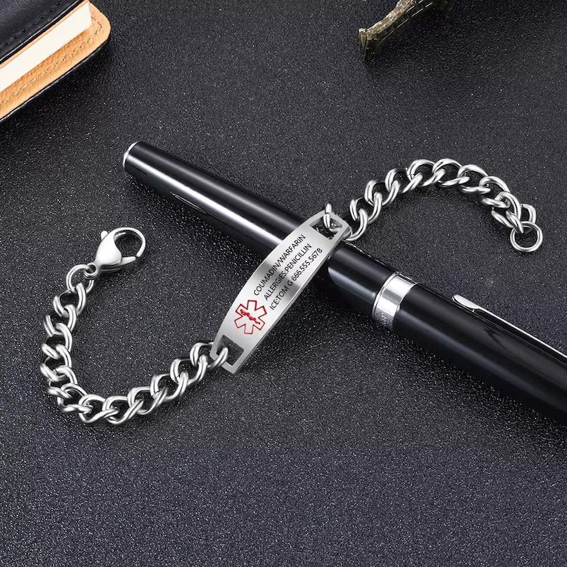 May include: A silver-colored medical ID bracelet with a chain link design. The rectangular tag displays the medical symbol and text: COUMADIN/WARFARIN, ALLERGIES: PENICILLIN, ICETOM G 666.555.5678. The bracelet is resting on a black pen.