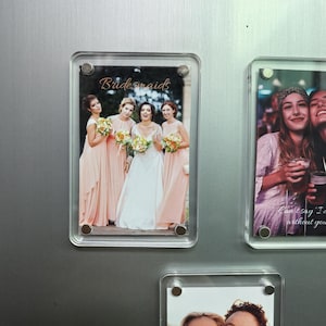 Könnte beinhalten: Ein klarer Acrylmagnetrahmen mit einem Hochzeitsfoto. Das Foto zeigt eine Braut in einem weißen Kleid mit Brautjungfern in pfirsichfarbenen Kleidern, die Blumensträuße halten. Das Wort "Bridesmaids" ist in Schreibschrift geschrieben.
