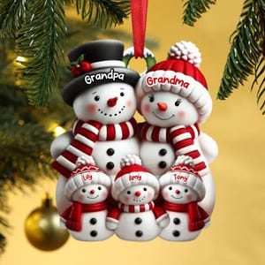 May include: A family of snowmen Christmas ornament. The snowmen are wearing red and white scarves and hats. The ornament has the names "Grandpa", "Grandma", "Lily", "Amy", and "Tony" on the hats. The ornament is hanging from a red ribbon.