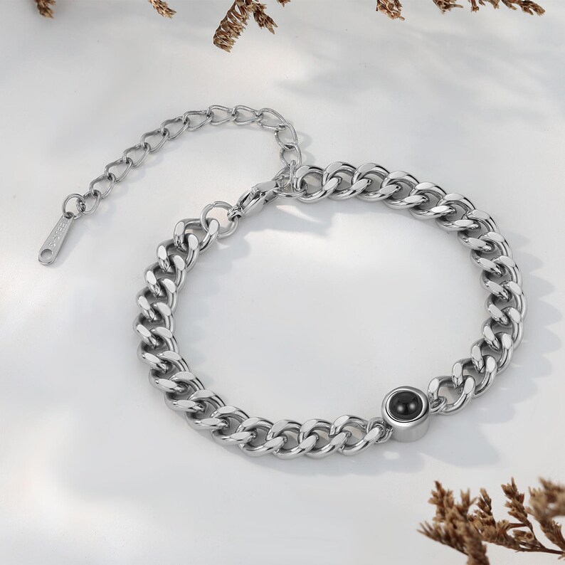 May include: A silver-colored chain bracelet with a large link design. The bracelet features a small, round, black stone set in a silver bezel. The bracelet has an adjustable chain for a custom fit. The jewelry is displayed on a white surface.