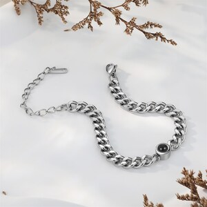 May include: A silver-colored chain bracelet with a large link design. The bracelet features a small black circular accent and a clasp closure. The bracelet is displayed on a white surface with dried floral accents.