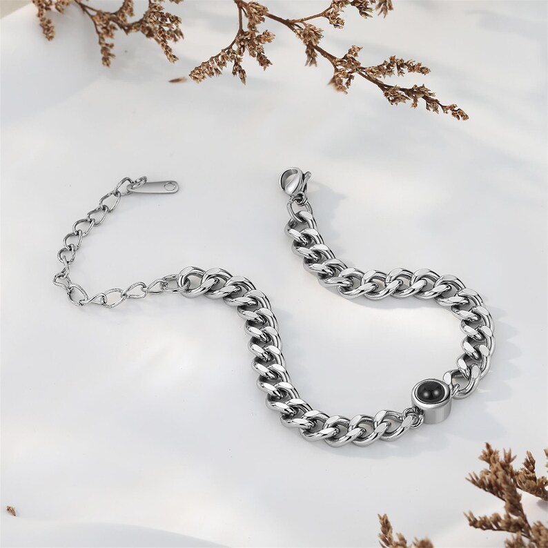 May include: A silver-colored chain bracelet with a large link design. The bracelet features a small black circular accent and a clasp closure. The bracelet is displayed on a white surface with dried floral accents.