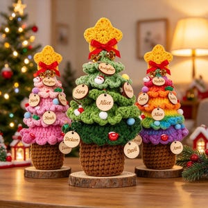 May include: Three crocheted Christmas trees on wooden bases. The left is pink, the center is green, and the right is rainbow-colored. Each features a yellow crocheted star, a red ribbon, small bells, and round wooden tags with names.