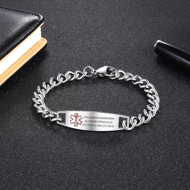May include: A silver-colored medical ID bracelet with a chain link design. The rectangular plate displays a red medical symbol and text: COUMADIN/WARFARIN, ALLERGIES: PENICILLIN, ICE: TOM G 666.555.5678. The bracelet is on a dark surface.