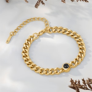 May include: A gold-colored chain bracelet with a black circular stone. The bracelet features a thick curb chain design and an adjustable clasp. The jewelry is displayed on a white surface.