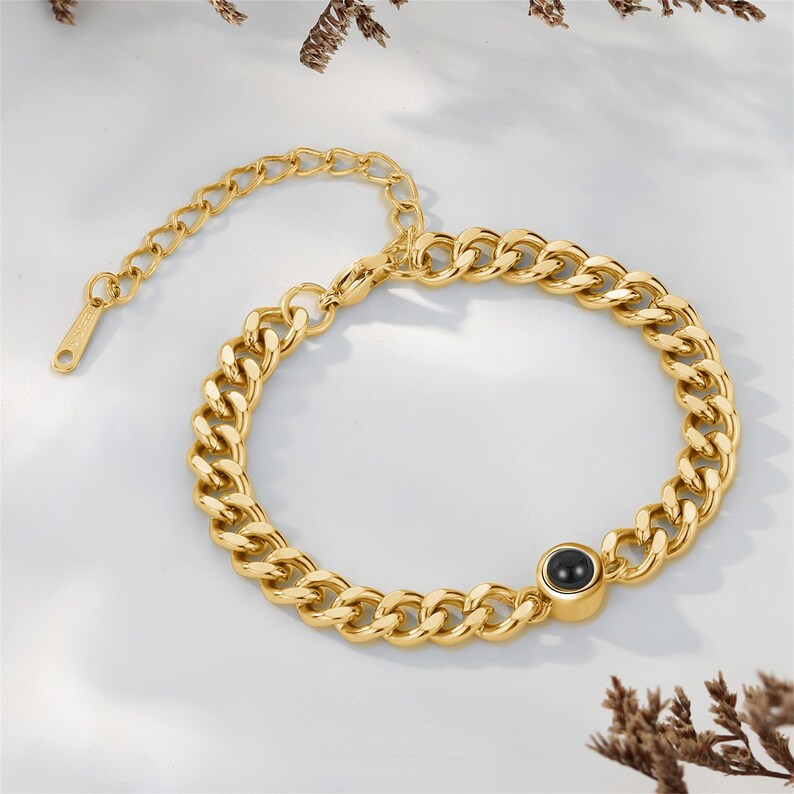 May include: A gold-colored chain bracelet with a black circular stone. The bracelet features a thick curb chain design and an adjustable clasp. The jewelry is displayed on a white surface.