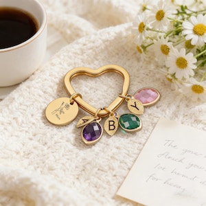 May include: Gold-colored heart-shaped keychain with a round charm engraved with a rose and the word "Mom". The keychain has several gemstone charms in purple, green, and pink, with the letters "A", "B", and "Y".