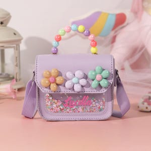 May include: A lavender crossbody bag with a beaded handle and flower embellishments. The bag features a clear front panel with colorful confetti and the name "Lucilla" in pink script. The bag has a lavender shoulder strap.