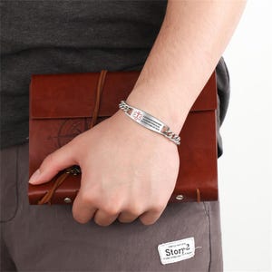 May include: A stainless steel medical ID bracelet with a curb chain design. The ID plate features a red medical symbol and printed text. The bracelet is shown on a wrist, with a brown leather journal in the background.