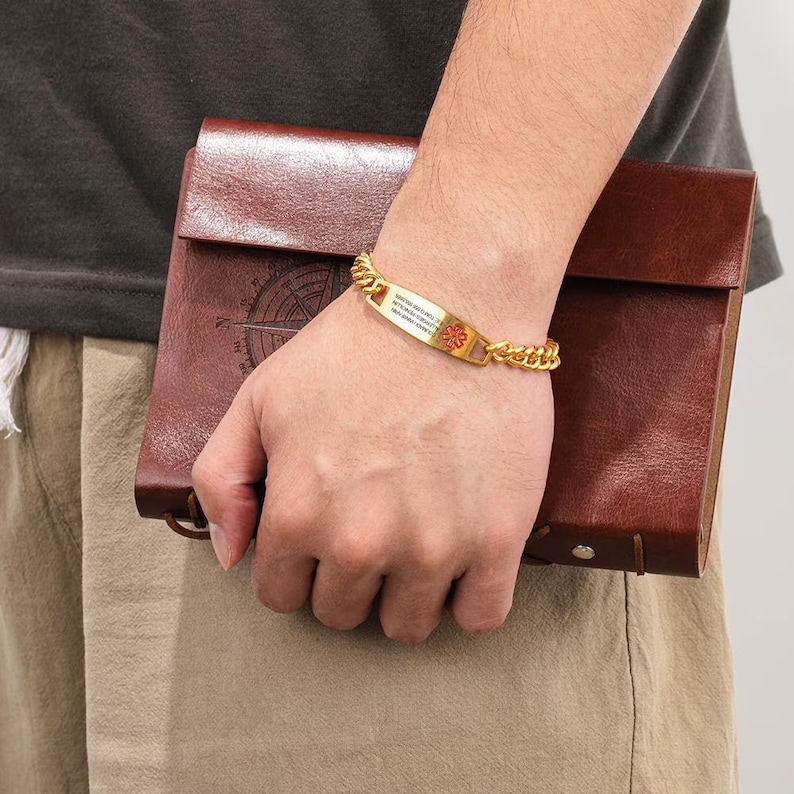 May include: A gold-colored medical ID bracelet with a curb chain design. The rectangular ID plate features a red medical symbol and printed text. The bracelet is worn on a wrist, and a brown leather notebook is held in the other hand.