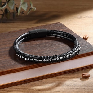 Può includere: Bracciale in pelle nera con chiusura magnetica. Il bracciale presenta un design intrecciato con dettagli in metallo color argento. Il bracciale è esposto su una superficie di legno, che ne mette in risalto la trama e il design.