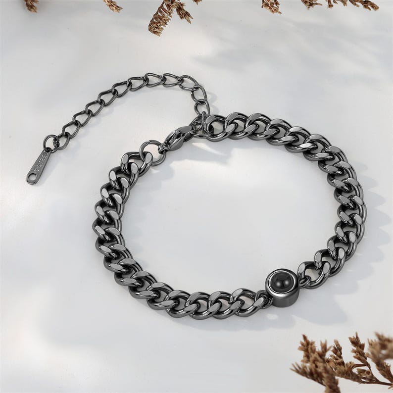 May include: A gunmetal-colored chain bracelet with a large curb chain design. The bracelet features a small, round, black stone accent and an adjustable chain closure. The jewelry is displayed on a white surface.