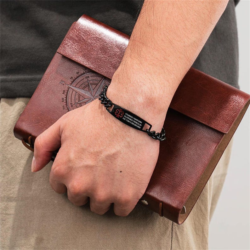 May include: Black medical ID bracelet with a chain link design. The bracelet has a rectangular plate with red medical symbols and text. The person is holding a brown leather journal with a compass rose design.