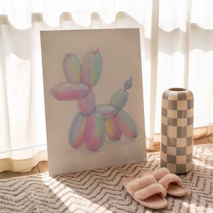 May include: A framed artwork depicts a balloon dog in pastel colors, including pink, blue, and yellow. A tall, checkered vase and a pair of pink slippers are also visible. The artwork is leaning against a wall near a window.