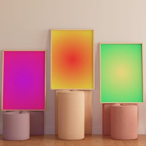 Op de afbeelding: Drie ingelijste gradiëntprints in gele, roze en groene kleuren, tentoongesteld op drie cilindrische sokkels in een moderne interieursetting.