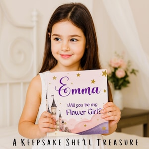 May include: A young girl holds a personalized book with the name "Emma" in purple script. The book cover features a castle illustration, gold stars, and the text "Will you be my Flower Girl?" and "A Keepsake She'll Treasure."