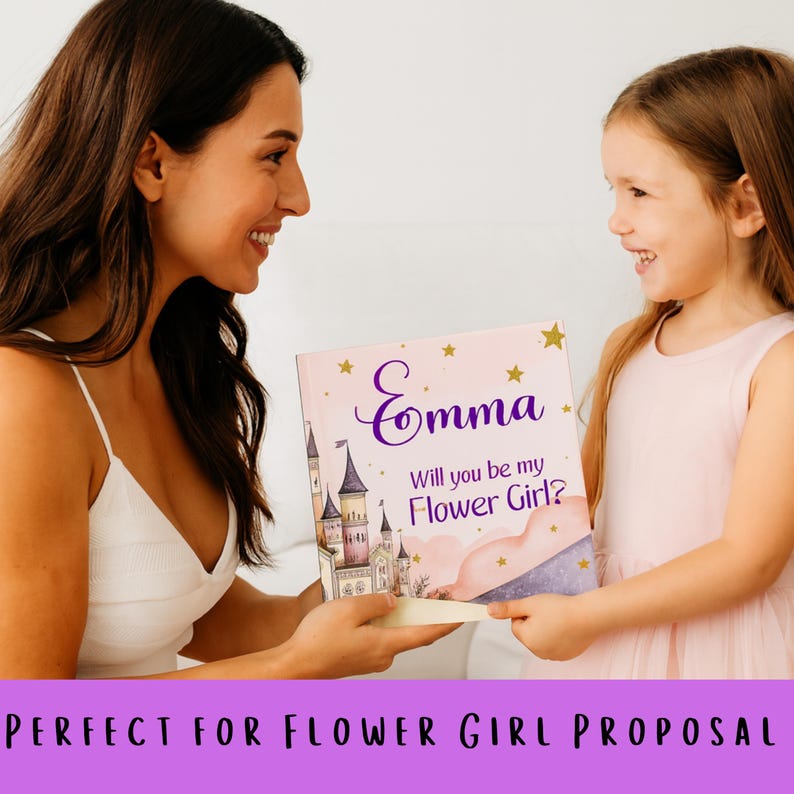 May include: A pink book with the name "Emma" in purple script and the text "Will you be my Flower Girl?" is held by two people. The book features a castle illustration and gold stars. The book is a flower girl proposal gift.