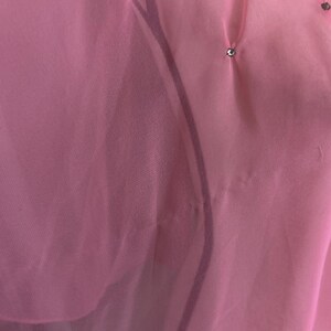 May include: Close-up of a sheer, light pink fabric with subtle pleats and small, sparkling embellishments. The fabric appears delicate and flowing, with a soft, translucent quality. The overall aesthetic is elegant and refined.