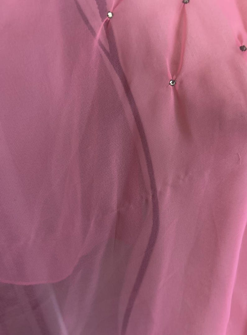 May include: Close-up of a sheer, light pink fabric with subtle pleats and small, sparkling embellishments. The fabric appears delicate and flowing, with a soft, translucent quality. The overall aesthetic is elegant and refined.
