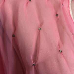 May include: Close-up of a sheer pink fabric with a textured appearance. Small, decorative embellishments are evenly spaced across the surface. The fabric is gathered at each embellishment, creating a subtle, dimensional effect.